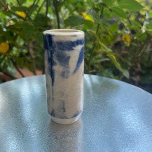 May include: A small, cylindrical ceramic vase with a white base and a cream-coloured body. The vase features a blue, marbled design. The vase is standing on a silver surface, with green foliage in the background.