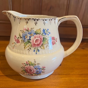 Puede incluir: Jarra de cerámica color crema con un diseño floral de flores rosas y azules. La jarra tiene un ribete dorado alrededor del borde y el asa. El asa es curva y la jarra tiene una base bulbosa.