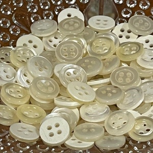 Op de afbeelding: Een grote groep witte plastic knopen met elk vier gaten. De knopen zijn in een willekeurig patroon gerangschikt en zijn allemaal even groot.