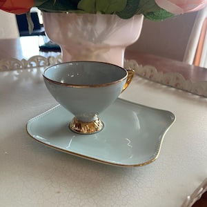 Può includere: Una tazza e un piattino azzurro con finiture dorate. La tazza ha un manico dorato e poggia su un piattino abbinato. Il set è su un vassoio bianco, con una composizione floreale sullo sfondo.