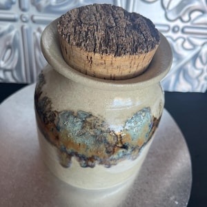 May include: A handmade ceramic jar with a cork stopper. The jar is off-white with a band of blue and brown glaze. The cork stopper is a dark brown colour. The jar is sitting on a silver surface.