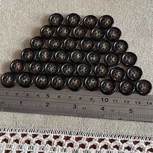 May include: A pile of 40 black buttons with four holes each. The buttons are arranged in a triangular shape on a beige fabric surface. A ruler is placed at the bottom of the image with markings in inches.