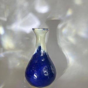 May include: A ceramic vase with a bulbous base and a narrow neck, featuring a glossy finish. The lower portion of the vase is a deep, vibrant blue, while the upper neck transitions into a creamy off-white colour with subtle blue speckling.