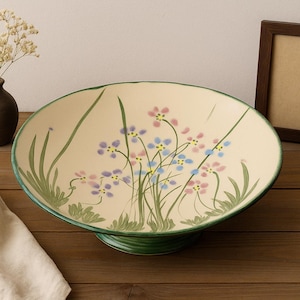 May include: A cream-colored ceramic serving bowl with a green rim and base, decorated with painted flowers in pink, blue, and purple. A small vase with dried flowers and a picture frame are also present.