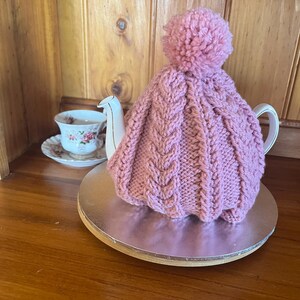 May include: A pink knitted tea cozy with a cable knit design and a large pom-pom on top. The cozy is covering a white teapot, sitting on a silver-colored round surface. A teacup and saucer with a floral design are visible in the background.