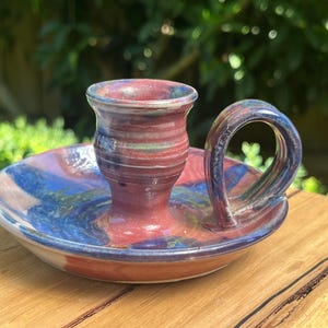 May include: A handcrafted ceramic candle holder with a loop handle. The candle holder features a blend of red, blue, and green hues. The base is a shallow dish, and the handle is a curved loop. It rests on a wooden surface.