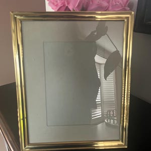 May include: A rectangular gold picture frame with a light-coloured mount. The frame is empty, reflecting the surroundings. Pink roses are visible above the frame. The frame is on a dark surface.