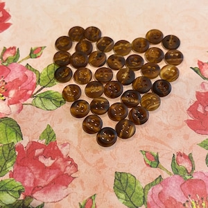 May include: A heart-shaped arrangement of brown, four-hole buttons with a marbled pattern. The buttons are arranged on a pink and white floral background.