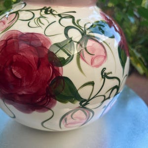 May include: A round ceramic vase with a cream-coloured base, featuring hand-painted dark red roses and light pink rosebuds with green stems and leaves. The vase has a smooth, glossy finish and a solid light pink rim around the circular opening.