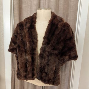 May include: A dark brown fur stole with short sleeves. The vintage wrap is displayed on a mannequin, showcasing its texture and shape. The fur appears soft and luxurious, perfect for adding warmth and style.