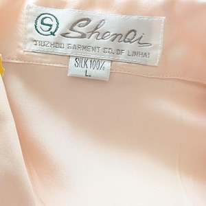 May include: A close-up of a light peach silk garment. The label reads "Shenli" with the text "TUZHOU GARMENT CO. OF LINHAI" and "SILK 100%" with a size "L". The fabric is smooth, possibly a blouse or shirt.