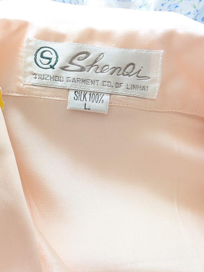 May include: A close-up of a light peach silk garment. The label reads "Shenli" with the text "TUZHOU GARMENT CO. OF LINHAI" and "SILK 100%" with a size "L". The fabric is smooth, possibly a blouse or shirt.