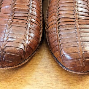 May include: Close-up of two brown leather shoes. The shoes have a woven design, with strips of leather interlaced to create a textured surface. The shoes appear to be well-worn, with visible creases and wear marks.