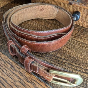 May include: A brown leather belt with a brass buckle is coiled on a wooden surface. The belt shows signs of wear and tear, with visible stitching and a worn appearance. The buckle is rectangular and has a shiny, golden finish.
