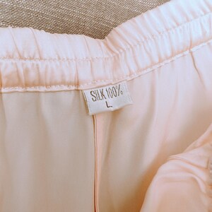 May include: Close-up of light pink silk trousers. The waistband is elasticated. A tag reads "SILK 100%" and "L". The fabric has a soft, flowing appearance.