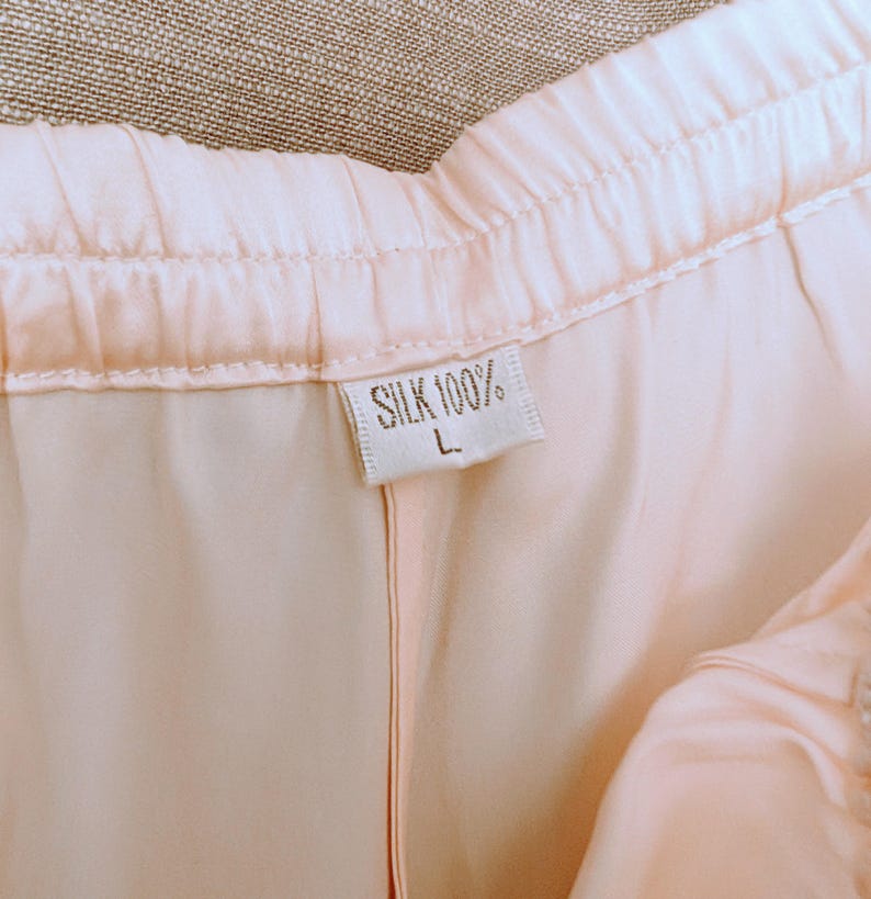 May include: Close-up of light pink silk trousers. The waistband is elasticated. A tag reads "SILK 100%" and "L". The fabric has a soft, flowing appearance.