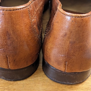 May include: Close-up of two brown leather loafers. The shoes have a textured surface and a dark brown sole. The leather shows signs of wear, adding to the vintage aesthetic. The image focuses on the heels of the shoes.