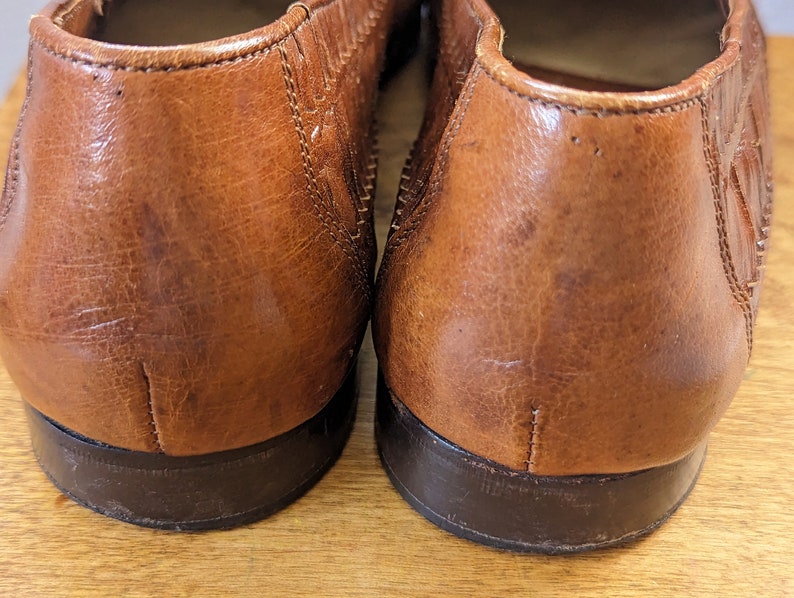 May include: Close-up of two brown leather loafers. The shoes have a textured surface and a dark brown sole. The leather shows signs of wear, adding to the vintage aesthetic. The image focuses on the heels of the shoes.