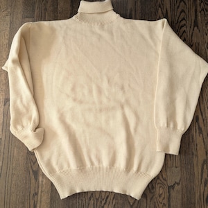 May include: Cream-colored turtleneck sweater. The sweater has long sleeves with ribbed cuffs and a ribbed hem. The turtleneck is also ribbed. The sweater is made of a soft, knitted material.