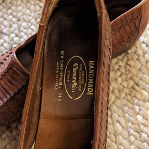 May include: Close-up of a pair of brown leather shoes with a woven design. The shoes are labeled "Churchills Shoes, New York-Beverly Hills, Made in Italy" and "Handmade". The shoes are on a woven surface.