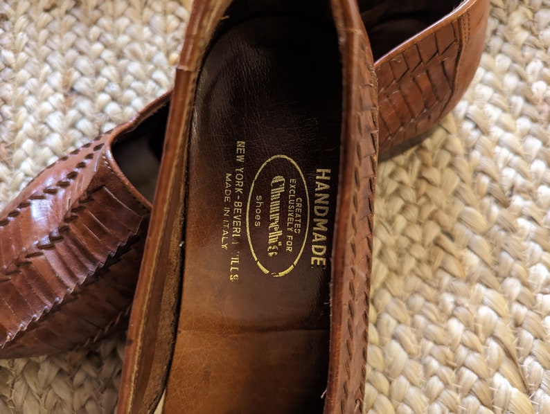 May include: Close-up of a pair of brown leather shoes with a woven design. The shoes are labeled "Churchills Shoes, New York-Beverly Hills, Made in Italy" and "Handmade". The shoes are on a woven surface.