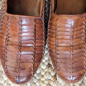 May include: Close-up of two brown leather shoes with a woven design. The shoes have a square toe and a flat sole. The leather has a rich, warm tone, and the woven pattern adds texture and visual interest. The shoes are likely sandals or flats.