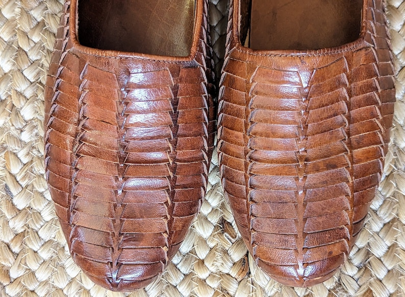 May include: Close-up of two brown leather shoes with a woven design. The shoes have a square toe and a flat sole. The leather has a rich, warm tone, and the woven pattern adds texture and visual interest. The shoes are likely sandals or flats.