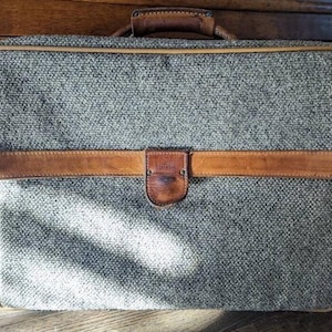 May include: A vintage gray tweed suitcase with a brown leather handle and trim. The suitcase has a small leather patch on the front.