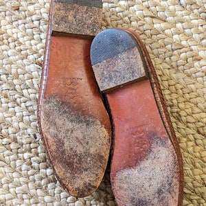 May include: A pair of well-worn, brown leather shoes, soles facing up. The soles show wear and tear, with visible dirt and scuff marks. The shoes have a woven texture on the upper part. The heels are made of a darker material.