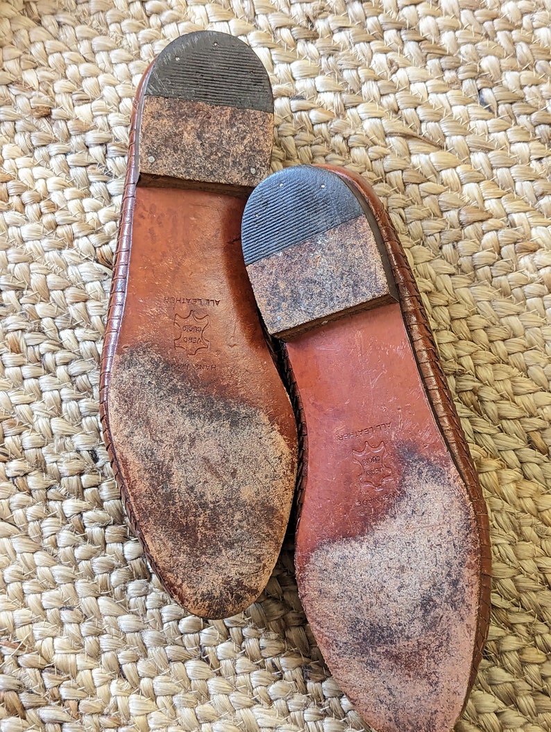 May include: A pair of well-worn, brown leather shoes, soles facing up. The soles show wear and tear, with visible dirt and scuff marks. The shoes have a woven texture on the upper part. The heels are made of a darker material.