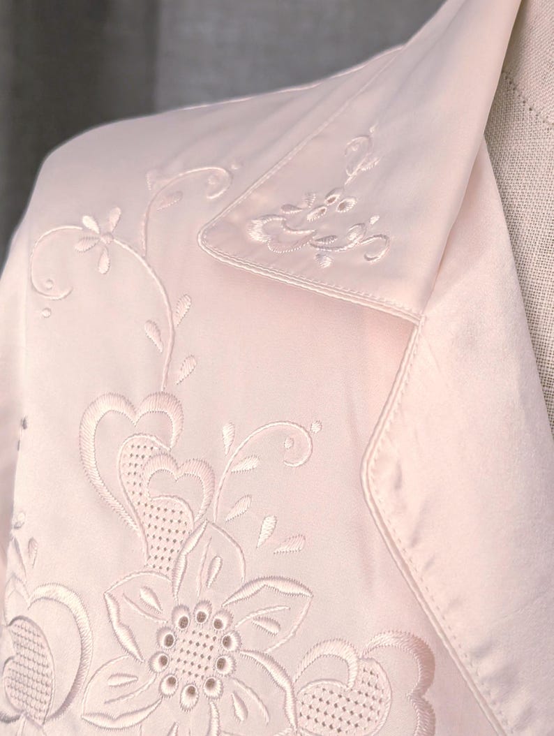 May include: Pale pink satin jacket with detailed floral embroidery. The jacket has a notched collar and embroidered flowers and leaves. The embroidery is in a matching pale pink thread, creating a delicate and elegant appearance.