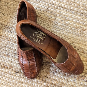 May include: A pair of brown leather flats with a woven design. The shoes have a rounded toe and a low heel. The interior of the shoe is stamped with "HANDMADE" and "Church's Shoes, New York-Beverly Hills, Made in Italy."