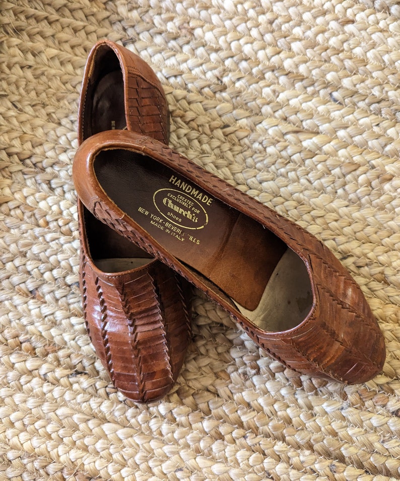 May include: A pair of brown leather flats with a woven design. The shoes have a rounded toe and a low heel. The interior of the shoe is stamped with "HANDMADE" and "Church's Shoes, New York-Beverly Hills, Made in Italy."