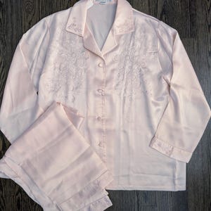 May include: A pale pink satin pajama set. The long-sleeved top has floral embroidery on the front and cuffs, with a button-down closure. The matching trousers are folded and have embroidered details at the hem. The set is on a dark wood surface.