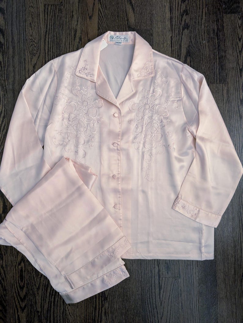 May include: A pale pink satin pajama set. The long-sleeved top has floral embroidery on the front and cuffs, with a button-down closure. The matching trousers are folded and have embroidered details at the hem. The set is on a dark wood surface.