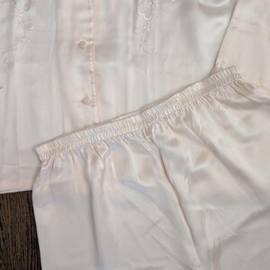 May include: A light peach-coloured pajama set. The top features embroidered floral detailing and buttons down the front. The pajama trousers have an elastic waistband. The set is made of a silky, smooth material.