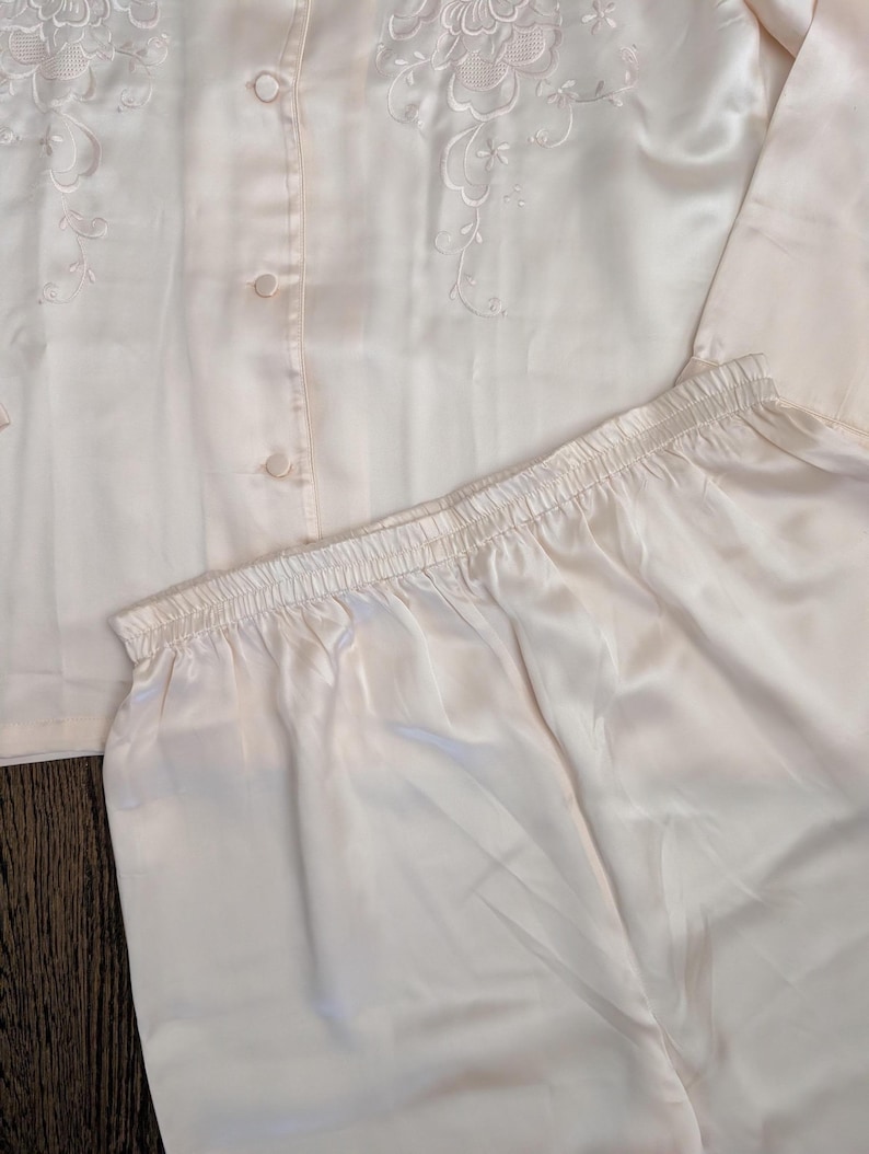 May include: A light peach-coloured pajama set. The top features embroidered floral detailing and buttons down the front. The pajama trousers have an elastic waistband. The set is made of a silky, smooth material.
