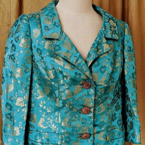 Op de afbeelding: Teal blazer met gouden bloemenpatroon. De jas heeft een reverskraag, drie bruine knopen en twee voorzakken. De stof heeft een subtiele glans en het bloemmotief is gedetailleerd.