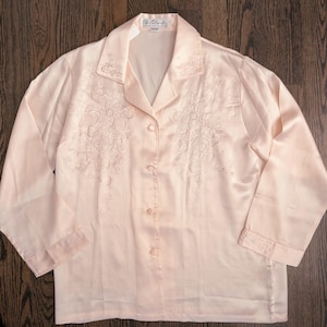 May include: A pale pink satin pajama top with floral embroidery. The long-sleeved top has a collared neckline and button-down front. The cuffs and collar are also embroidered. The top is made of a shiny, silky material.
