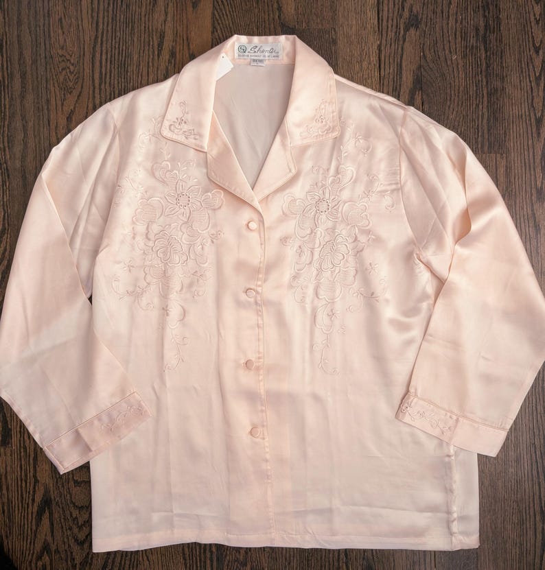 May include: A pale pink satin pajama top with floral embroidery. The long-sleeved top has a collared neckline and button-down front. The cuffs and collar are also embroidered. The top is made of a shiny, silky material.