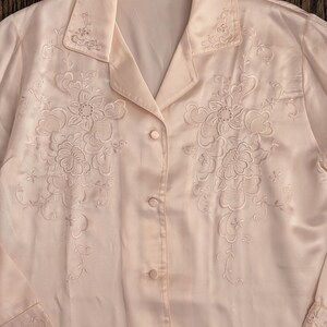 May include: Pale pink satin blouse with floral embroidery. The long-sleeved shirt features a collared neckline and button-down front. Delicate floral embroidery adorns the front and collar. The label reads "Shenli".