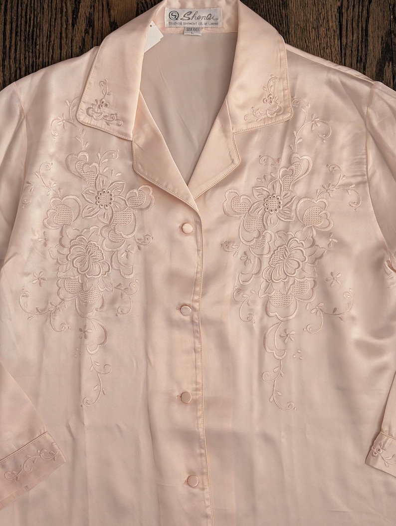 May include: Pale pink satin blouse with floral embroidery. The long-sleeved shirt features a collared neckline and button-down front. Delicate floral embroidery adorns the front and collar. The label reads "Shenli".