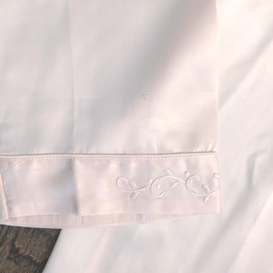 May include: Close-up of a white garment with a decorative embroidered floral design along the hem. The fabric appears to be a soft, smooth material. The embroidery is delicate and detailed.