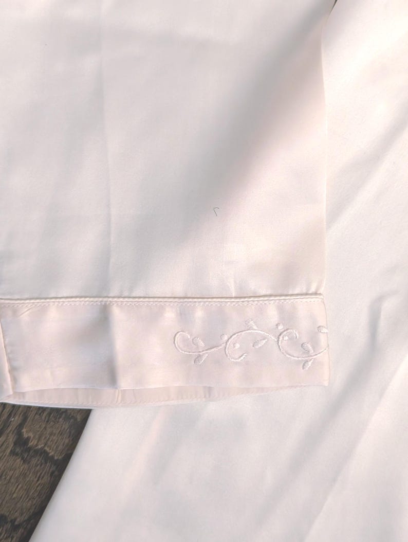 May include: Close-up of a white garment with a decorative embroidered floral design along the hem. The fabric appears to be a soft, smooth material. The embroidery is delicate and detailed.