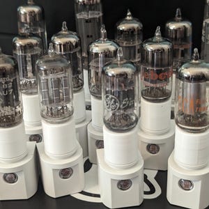 May include: A collection of vintage vacuum tubes with silver and black metal casings. The tubes are mounted on white bases with a red button on the bottom. The tubes have various labels, including "GE", "ECC82", "AU7", "WEBCOR", and "PATENTS INTERNATIONAL".