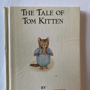 Puede incluir: Una copia vintage de "The Tale of Tom Kitten" de Beatrix Potter. La portada presenta una ilustración de un gato con un atuendo azul. El título y el nombre del autor están impresos en negro, con el logotipo de la editorial en la parte inferior.