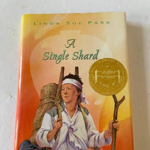 Puede incluir: Una portada de libro con fondo amarillo y naranja. El título "A Single Shard" está escrito en negro. El autor "Linda Sue Park" está escrito en negro. Una persona joven con una mochila y un bastón de senderismo está en la portada. El libro tiene una pegatina de premio dorada.