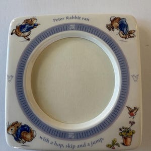 May include: A square ceramic picture frame featuring illustrations of Peter Rabbit in a blue jacket. The frame has a circular center opening, a blue border, and the text "Peter Rabbit ran with a hop, skip and a jump." Small birds and a potted plant are also depicted.