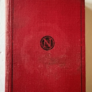 Pode incluir: Um livro vintage com capa vermelha. A capa tem uma aparência texturizada e uma borda preta. Um emblema circular com a letra "N" está no centro. As bordas do livro mostram sinais de desgaste.
