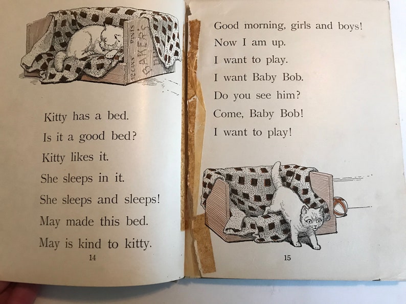 May include: A black and white illustration of a cat sleeping in a bed. The cat is white with black markings. The bed is made of wood and has a checkered blanket. The text "Kitty has a bed. Is it a good bed? Kitty likes it. She sleeps in it. She sleeps and sleeps! May made this bed. May is kind to kitty." is printed below the image. The page number "14" is printed at the bottom of the page.